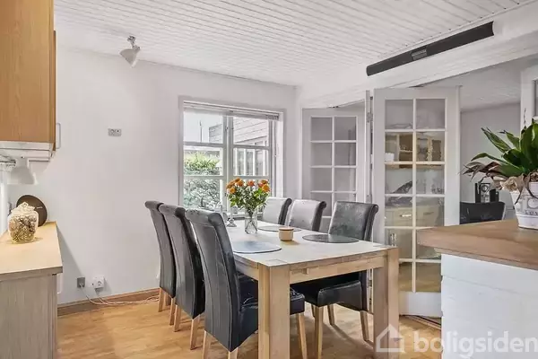 Spisebord omgivet af sorte stole står i et lyst rum med trælaminatgulv. En buket farverige blomster står på bordet, nær et vindue med udsigt til grønne planter.