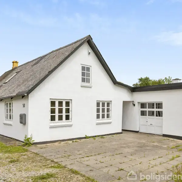 Det hvide hus med stråtag står på en gårdsplads med fliser. Det er omgivet af grønne træer og buske under en blå himmel.
