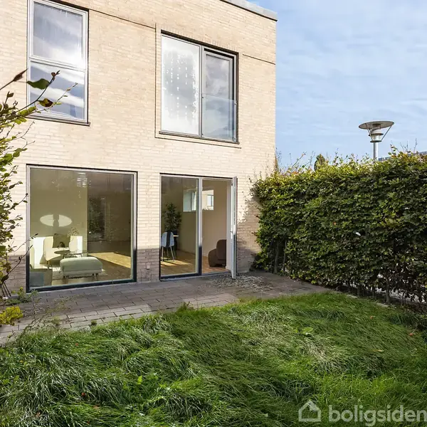 Toetagers hus med store vinduer vender ud mod en velplejet have med grønne planter. Tætklippede hække omkranser området, og en græsplæne ligger foran husets terrasse.
