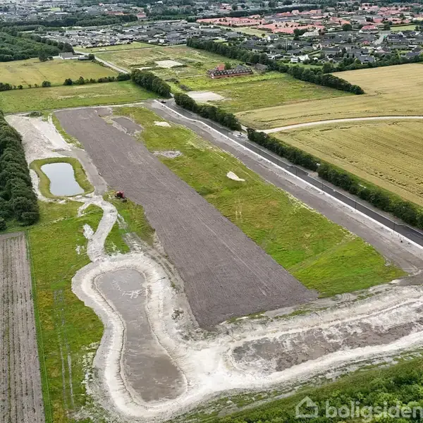 En stor byggeplads med jord og grus, set fra luften, omgivet af marker og træer. I baggrunden ses en by med mange huse.