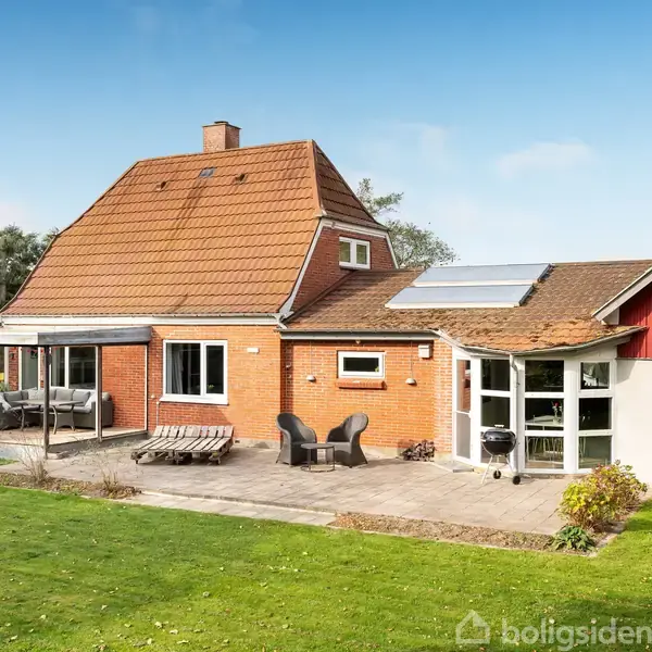 Rødt hus står i solrige omgivelser. Der er en terrasse med havemøbler foran huset, og en velplejet græsplæne omgiver ejendommen. Huset har solpaneler på taget.