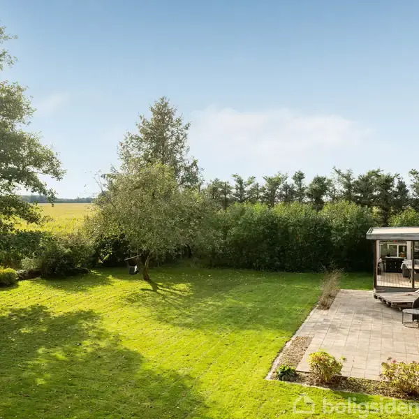 En grøn græsplæne med et centralt træ strækker sig foran en terrasse med havemøbler, omgivet af buske og træer i baggrunden. Himlen er klar og blå.