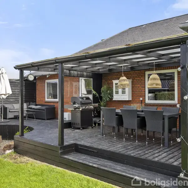 Overdækket terrasse med spisebord og stole står tæt på en grill, omgivet af murstenshus og træhavemøbler. Grøn græsplæne omkranser området, skaber en hyggelig udendørs atmosfære.