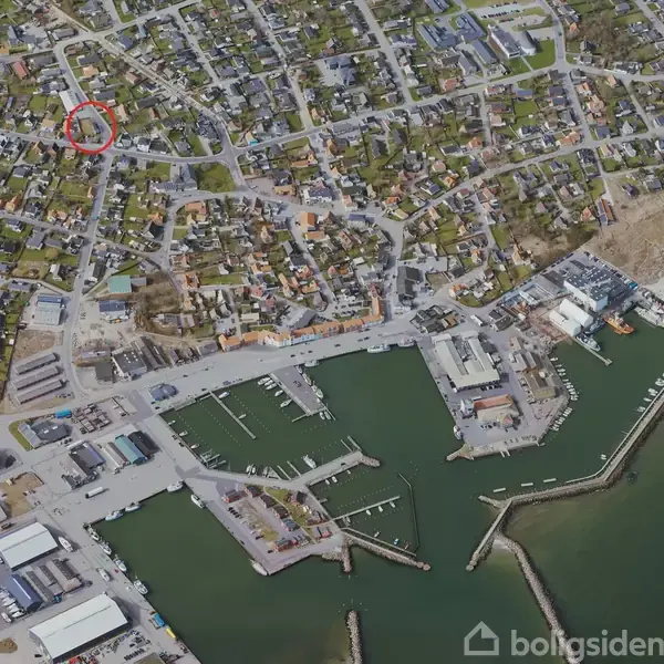 Byområde vises ovenfra med tætliggende huse og gader. En havn med flere kajanlæg og både ligger ved vandet i forgrunden. Et område er markeret med en rød cirkel.