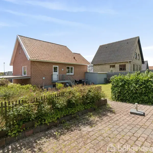Rødt murstenshus med skråt tag står i en velplejet have med en lille terrasse. En anden bygning ses i baggrunden ved siden af en hæk og brolagt gangsti.