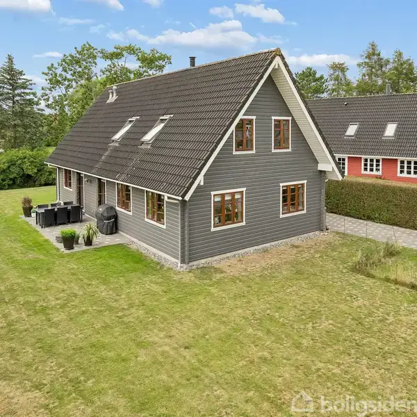 Gråt hus med rødt tag og hvide vinduer, står på en velplejet græsplæne. Til højre ses et tilsvarende hus i rødt. På terrassen står havemøbler og en grill.