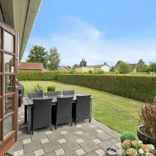 En udendørs terrasse med et spisebord og sorte stole står på fliser ved siden af et hus. I baggrunden ses en velplejet have med hække og græsplæne.