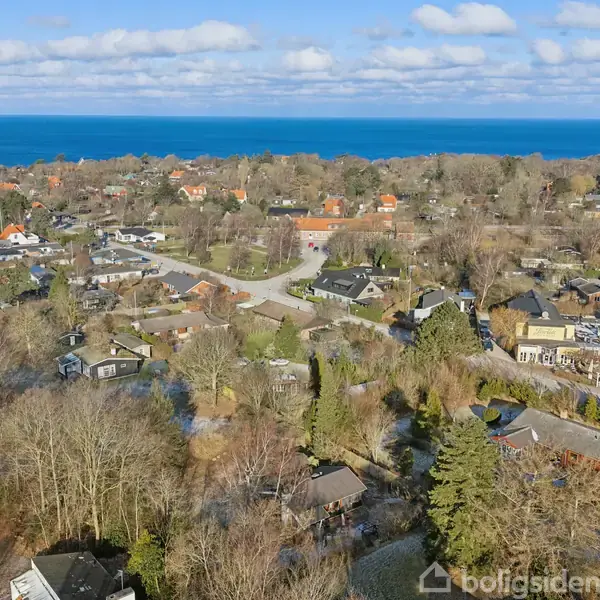 En by omgivet af træer, set fra luften, med mange huse spredt ud i landskabet. Havet ses i horisonten under en blå himmel med skyer.