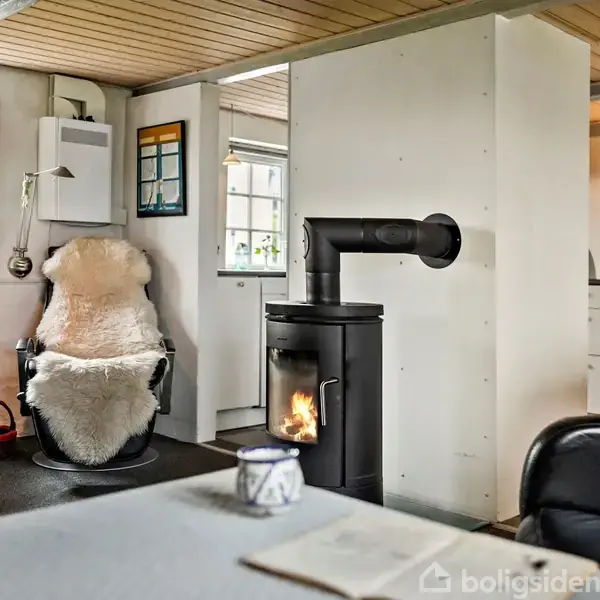 Brændeovn opvarmer stue med hyggelig atmosfære. Komfur står ved skillevæg; lænestol med fåreskind ved bord med blomster. Kaffe på bord ved forgrund. Interiøret er lyst og indbydende.