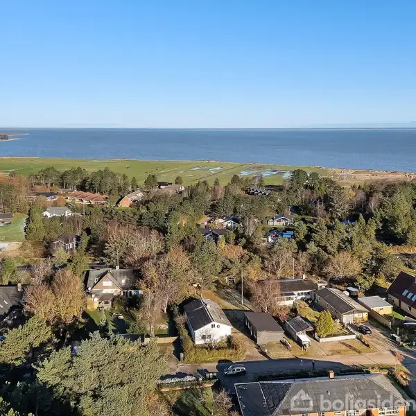 Beboelseshuse ligger spredt blandt træer tæt på en kystlinje med udsigt over havet. Grønt landskab og skov omkranser området under en klar blå himmel.