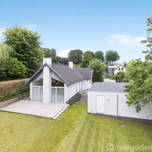 Et moderne hus står stille i en grøn have med stor terrasse. Omgivet af træer og hække, vises også en tilstødende garage.