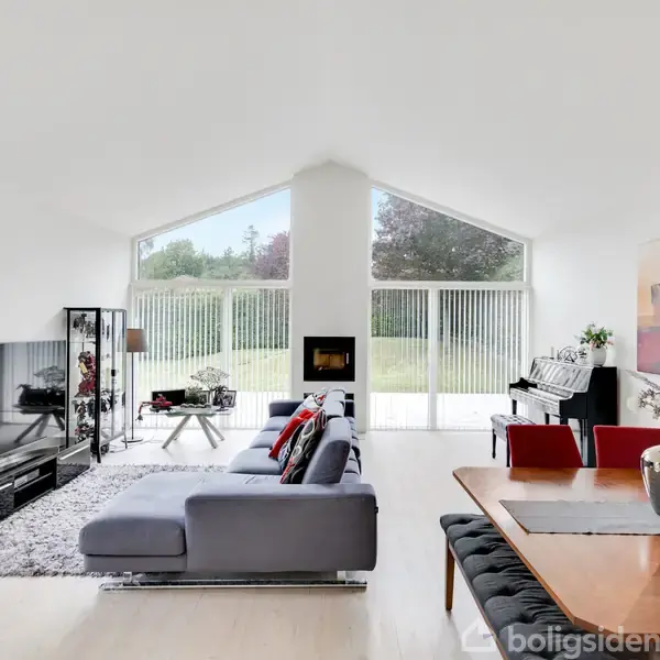 En stue med en grå sofa vender mod et stort fladskærms-tv. Store vinduer slipper lys ind. Der er også et spisebord, klaver, maleri på væggen og udsigt til haven.