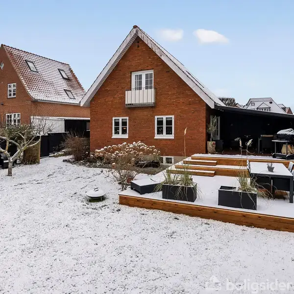 Rødt murstenshus med spidst tag står i en snedækket have. Træterrasse med planter og havemøbler, omgivet af sne. Naboer i baggrunden.