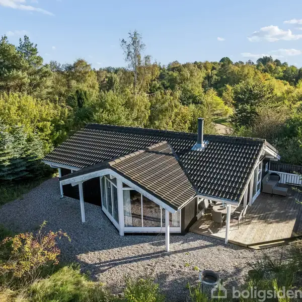 Et sommerhus med sorte teglstenstag omgivet af tætte, grønne træer. Huset har store vinduer og en træterrasse, der åbner ud mod en grusbelagt have.