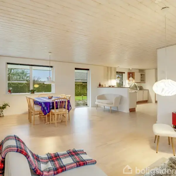 Sofa med tæppe står i et lyst stueområde; spisebord med stole i baggrunden tæt på store vinduer, åben forbindelse til et moderne køkken. Pejs med stole ved siden.