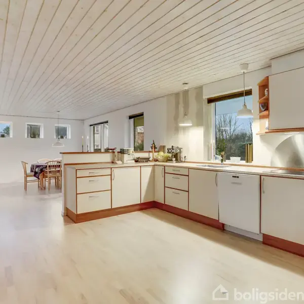 Køkken med hvide skabe og trækanter, arrangeret i en vinkel, åbner sig mod en spisestue. Lyse trægulve og hvide lofter skaber en lys atmosfære.