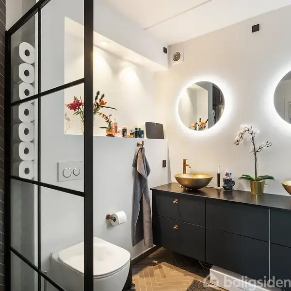 Toilet og håndvaske med to runde spejle i et moderne badeværelse. Håndklæder og toiletpapir er arrangeret ved siden af toilettet. Planter og småting står på sorte skabe med messingvaske.