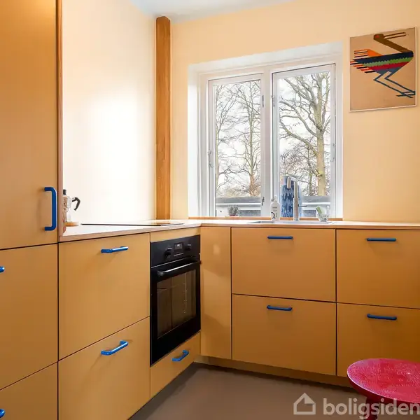 Et køkken med lysebrune skabe og blå håndtag. En ovn er indbygget i skabsvæggen. Vindue viser træer udenfor. En rød taburet står ved siden af vinduet. Et grafisk maleri hænger på væggen.