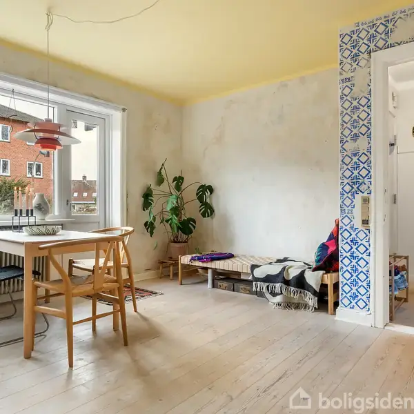 Et spisebord med stole står i et lyst rum med store vinduer. Til højre er en dør med blå-hvide fliser. En grøn plante og dekorationer ses i hjørnet.