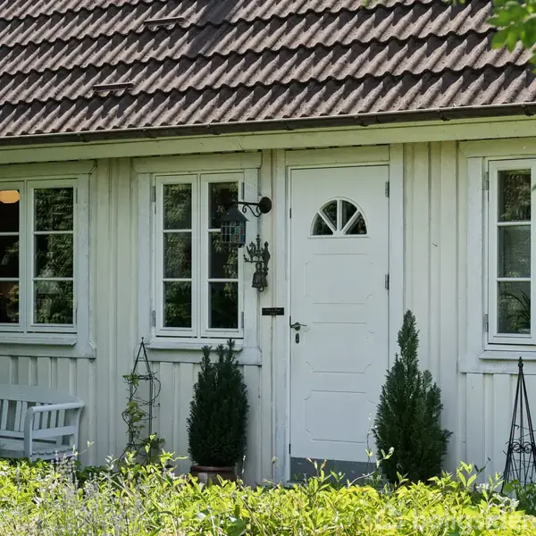 Hvidt træhus med en hvid dør og to vinduer, flankeret af små buske. En hvid bænk står til venstre. Grønne planter omgiver huset i en frodig have.