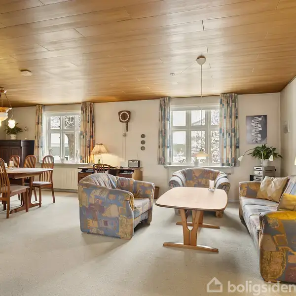 Stue med farverige møbler arrangeret omkring et sofabord; spisebord med stole i baggrunden. Rummet har store vinduer med gardiner, træloft og diverse lamper og dekorationer.