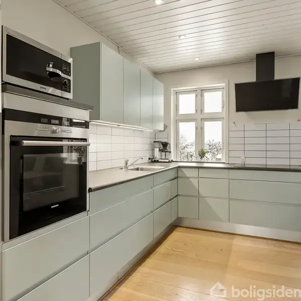 Køkken med lyse blågrønne skabe og stål apparater, inklusive indbygget ovn og mikroovn. Rummet har trægulv, flisevægge og store vinduer mod højre, der giver naturligt lys.