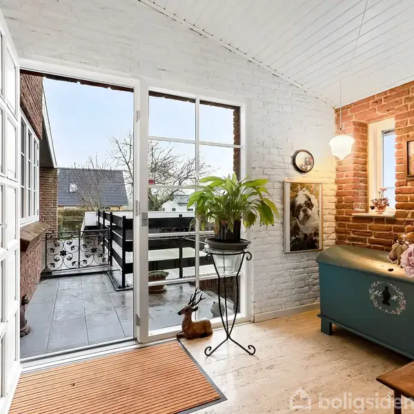 En entre med hvide vægge og store vinduer. En plante står på et metalstativ, mens en blå kiste dekoreret med blomster står ved en murstensvæg. En dør fører ud til en balkon.