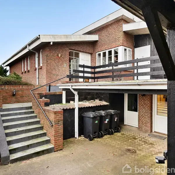 En murstensvilla med trappe til hovedindgangen, omgiven af beplantning. Der er en terrasse med gelænder og skraldespande nedenfor. Klare vinduer vender ud mod en flisebelagt gårdsplads.