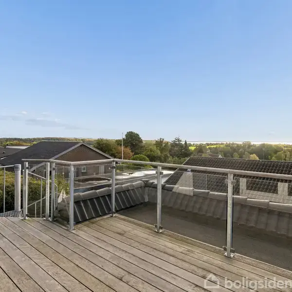 En træterrasse med glasrækværk står stille og tilbyder udsigt over hustage og grønne områder i en solrig landsbyzone.