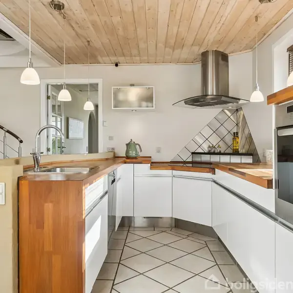 Moderne køkken med hvide skabe arrangeret i en vinkel. Over ovnen og kogepladen hænger en køkkenventilator. Der er et bord med vask, og en vindeltrappe i baggrunden.