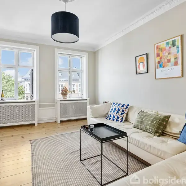 Stue med lys sofa, pyntet med farvede puder, stående ved siden af et rektangulært sofabord. En elektrisk guitar står ved radiatoren. Vægge har kunstværker. Store vinduer med udsigt til træer.