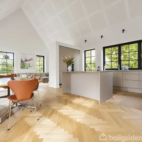 Køkken-alrum med moderne møblering; spisebord med orange stole i forgrunden; køkkenø centralt; store vinduer, der slipper naturligt lys ind; træparketgulv; hvidmalede vægge med højt loft.
