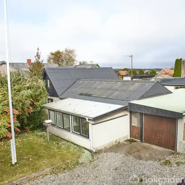 En enkeltstående bygning med grå tag og garagedør ligger i et grønt område omgivet af træer og buske. En grusvej og flagstang ses foran bygningen.