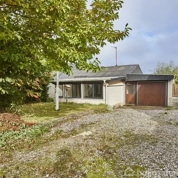 Et enfamiliehus med enkelt etplansdesign står stille på en grusbelagt indkørsel. Baggrunden er en frodig have med træer under en lys, let overskyet himmel.