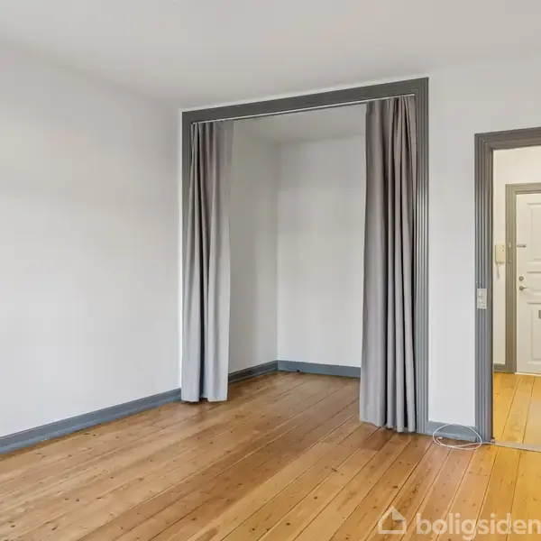 Værelse med trægulv, grå gardiner adskiller en alkove fra hovedområdet, dør mod en gang med lys trægulv. Væggene er hvide, og der er en enkel indretning.