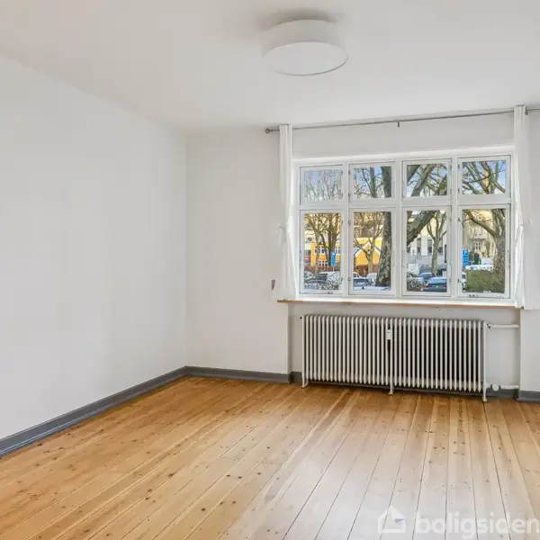 Et tomt værelse med trægulv, lyst malede vægge og en radiator under et stort vindue, der slipper dagslys ind. Udenfor ses træer og bygninger gennem vinduet.