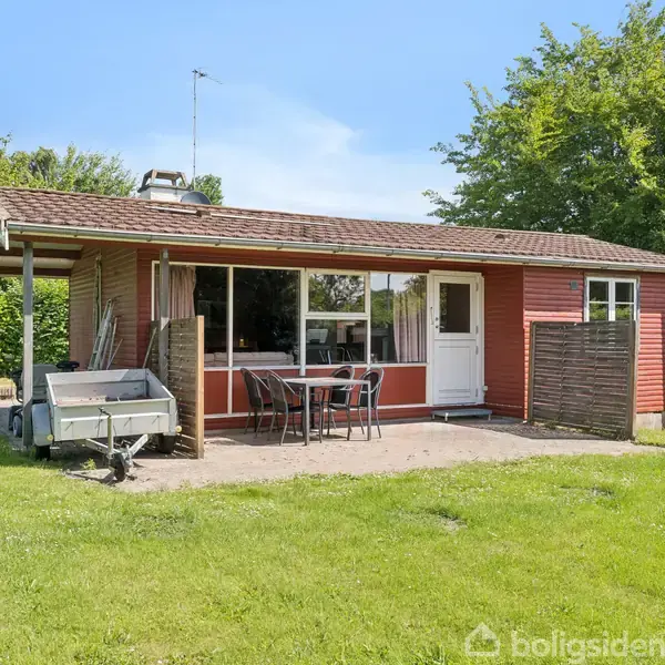 Rødt sommerhus med hvide døre og vinduer, omkranset af grønne træer og græsplæne. En terrasse foran har to stole og et bord. En trailer står ved siden af huset.