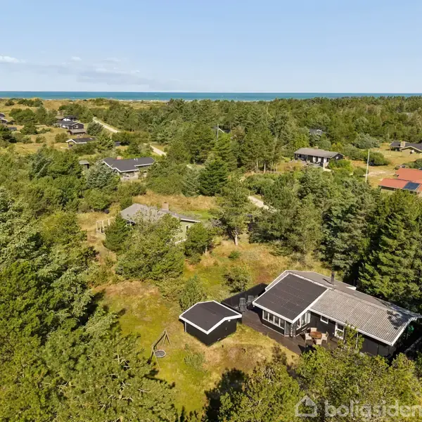 Et sommerhus omgivet af frodig skov og andre huse i et kystnært naturområde. I baggrunden ses havet.