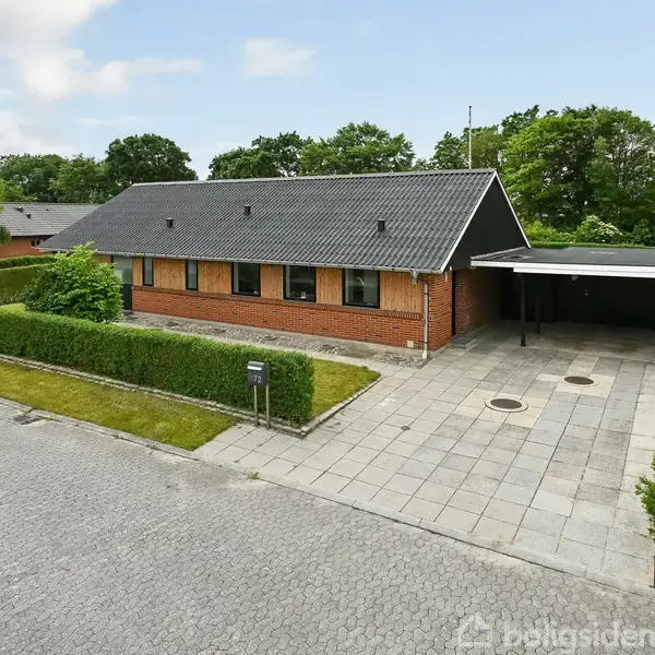 En enkel etplansrødstensvilla med sort tag står stille ved en flisebelagt indkørsel og carport. Grønt græs og hække omkranser huset i et villakvarter.
