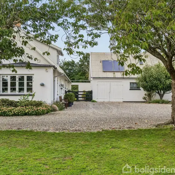 Hvidt hus med mørkt tag omgivet af træer i en grusbelagt gård. Til højre ses en tilbygning med solpaneler. Grøn græsplæne i forgrunden.