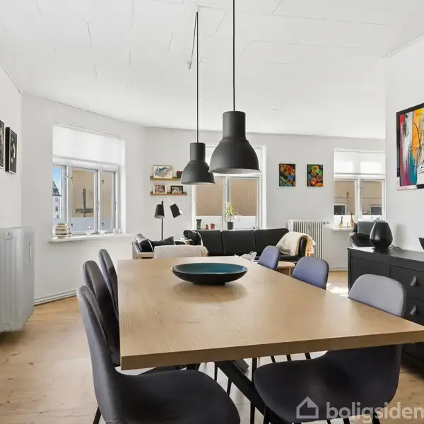 Spisebord med sorte stole, omgivet af moderne kunst på væggene i en lys spisestue med store vinduer og planteliv. En skænk står i hjørnet med udsmykning.
