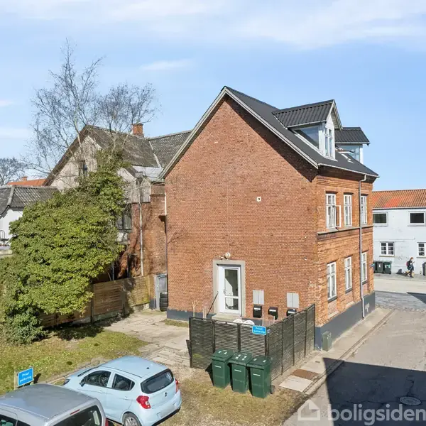 Rødt murstenshus står langs en stille gade. Flere skraldespande er placeret foran huset. I baggrunden ses parkerede biler og nabobygninger under en klar blå himmel.