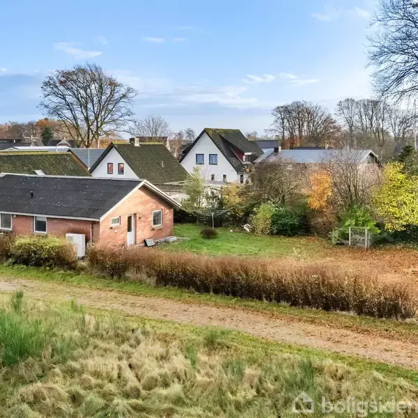 Et rødt murstenshus står stille i en landsby omgivet af grønne træer og buske. Der er flere huse i baggrunden på en klar dag med blå himmel.