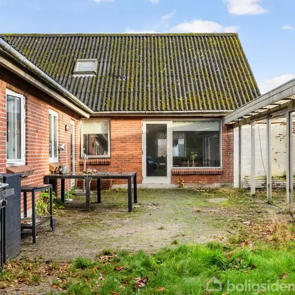 En udendørs gårdhave med et rødt murstenshus og grønt taget. Der er en grill og et bord med stole. Til højre ses en overdækket terrasse.