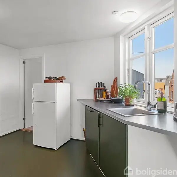 Køkken med hvid køleskab ved sidevæggen, grønne skabe og sort bordplade under et stort vindue. Diverse køkkenredskaber og planter på bordpladen. Sollys skinner ind.