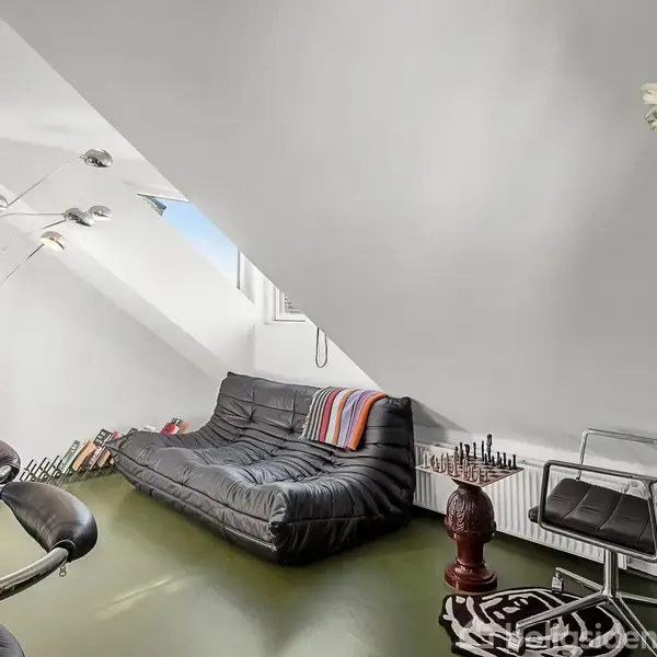 Sort lædersofa står op ad en skrå væg under et ovenlysvindue; en gulvlampe i metal lyser ved siden af en stol og et lille bord med skakbræt, i et moderne værelse.