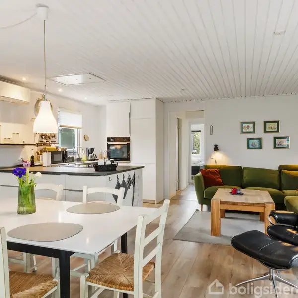 Stue med spisebord og stole i forgrunden, en sort lænestol ved siden af et grønt sofaarrangement. Køkkenet er i baggrunden med hvide skabe og hængende lamper. Blomster på bordet.