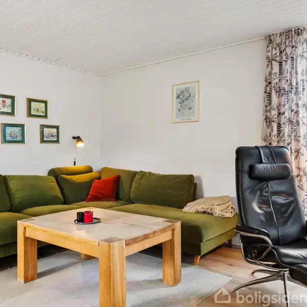 Grøn sofa med farverige puder står i stue, ledsaget af en sort lænestol og træbord. Væggene er dekoreret med billeder, mens vinduet har udsigt til en have.