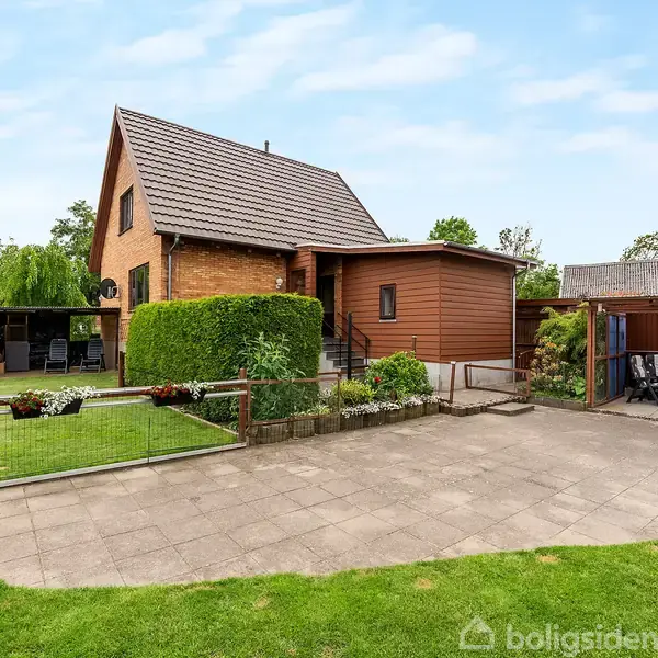 Hus i brun mursten med spidst tag, omgivet af velplejet have og terrasse. Til højre ses et åbent skur med overdækning og siddepladser. Græsplæne og flisebelagt areal foran.