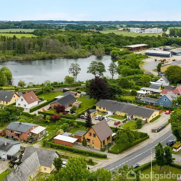 Flere huse ligger spredt langs en vej i et grønt, landligt område med udsigt til en stor sø bagved. Træer omgiver søen, og der er marker i baggrunden.